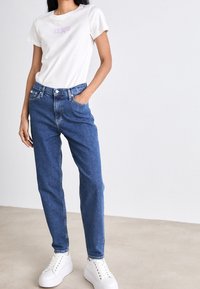 Bílý krátký rukáv tričko s fialovou grafikou, spárované s modrými rovnými džíny a bílými platformovými teniskami. Jednoduché pozadí.