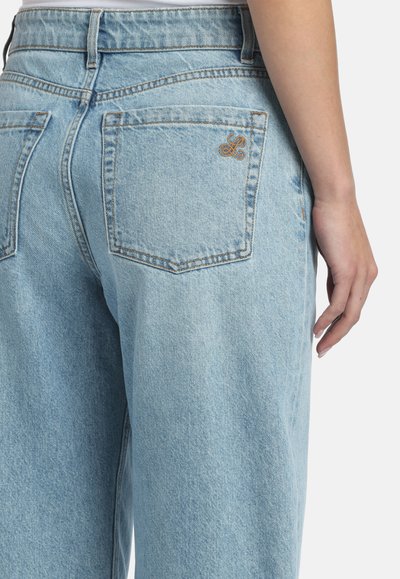 Svjetlo plave traperice na osobi, prikazujući stražnji džep s malim izvezenim logotipom i dio desne ruke.