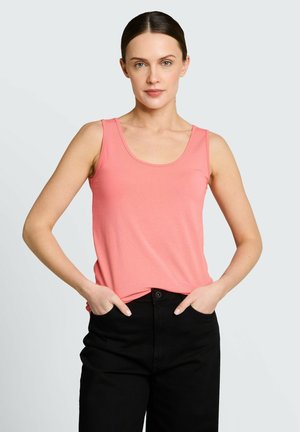 Kobieta o ciemnych włosach, ubrana w różowy top bez rękawów i czarne spodnie, stoi z rękami w kieszeniach na tle jasnoszarym.