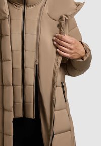 Béžový prošívaný kabát s vysokým límcem, dvěma kapsami na zip a prošívanou strukturou, se zdvojeným předním zipem a lesklými černými kovovými doplňky.