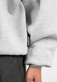 Šedá mikina s měkkou texturou, na rukávu s černou cedulkou s nápisem "PROHIBIT" a v kombinaci s tmavě šedými kalhotami.