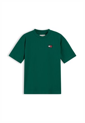 Jeune garçon souriant tenant un pull vert en maille torsadée, portant un t-shirt vert, un jean foncé et des baskets blanches, sur fond blanc.