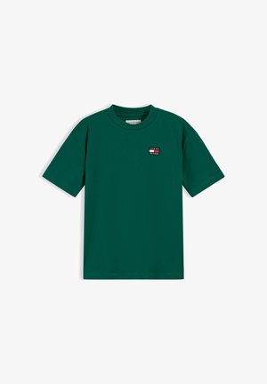 Jeune garçon souriant tenant un pull vert en maille torsadée, portant un t-shirt vert, un jean foncé et des baskets blanches, sur fond blanc.