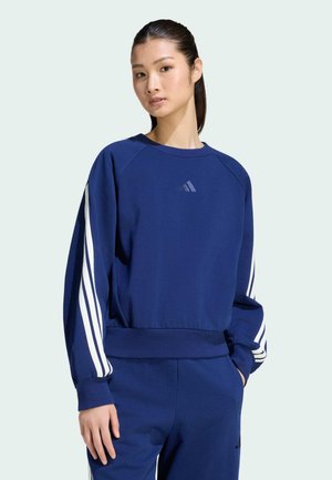 Ung kvinde med mørkt hår i en blå Adidas træningsdragt med hvide striber på ærmer og bukser, stående med den ene hånd i lommen.
