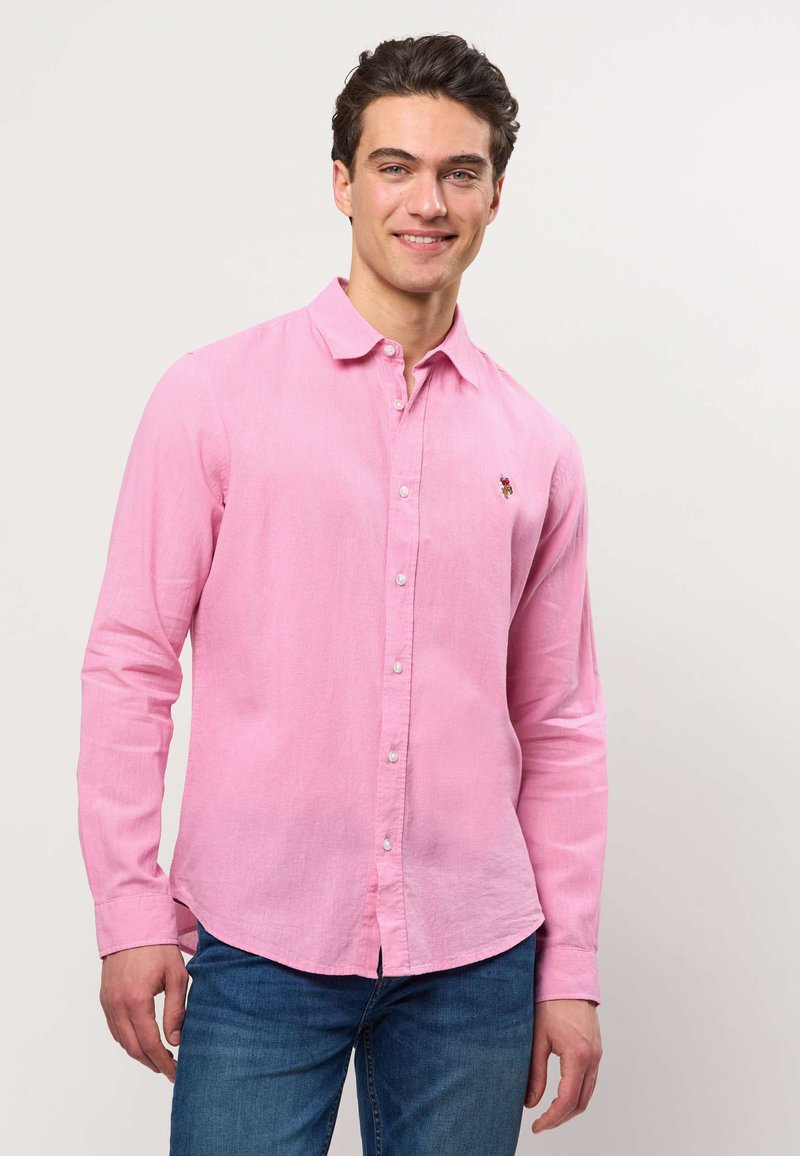 Homme souriant portant une chemise rose clair à manches longues boutonnée et un jean bleu, debout devant un fond clair uni.