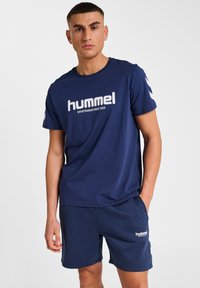 Dunkelblaues Sport-T-Shirt, kurze Ärmel, mit weißem "hummel"-Logo und gestreiften Akzenten auf den Schultern. Kombiniert mit dunkelblauen Shorts mit Logo.