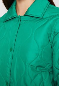 Nahaufnahme einer grünen gesteppte Jacke mit Druckknöpfen und Kragen, die von einer Person getragen wird, mit sichtbaren Stofftexturen und Nähdetails.