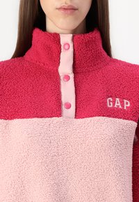 Fleece pullover in roze en lichtroze. Beschikt over een hoge kraag, drukknoopjes in lichtroze en een subtiel geborduurd "GAP" logo.