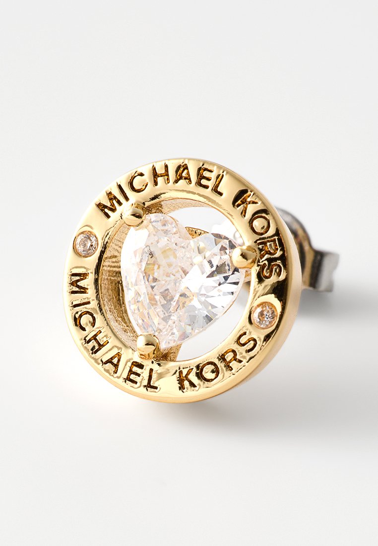 Pendiente de Stud en tono dorado con un centro de cristal transparente en forma de corazón, rodeado por una banda circular grabada con el texto "MICHAEL KORS".