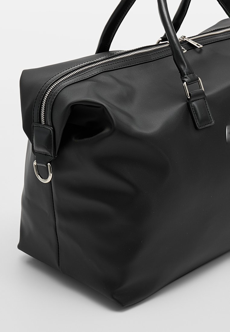 Bolso de material sintético negro texturizado con cierre de cremallera, dos asas y un adorno metálico. Forma suave y redondeada con diseño minimalista.