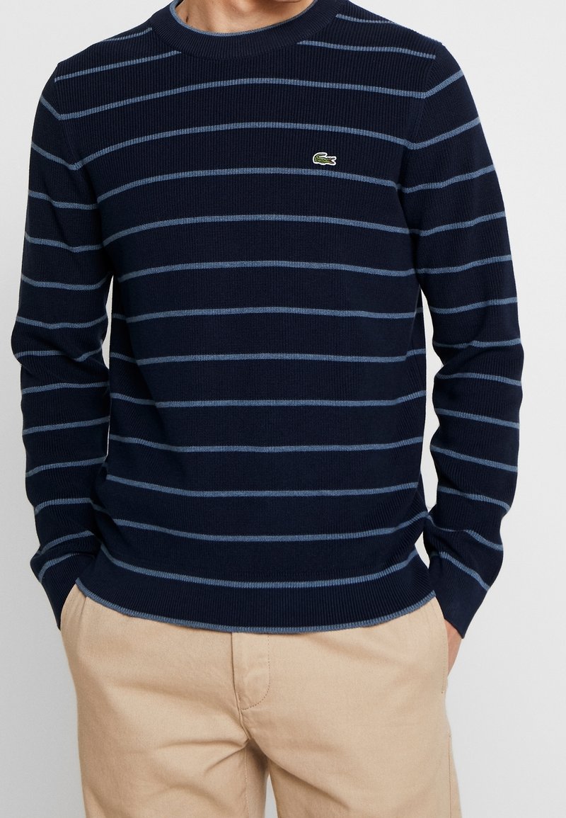 Homme portant un pull bleu marine avec des rayures horizontales bleu clair et un petit logo crocodile vert, associé à un pantalon beige, les mains dans les poches.