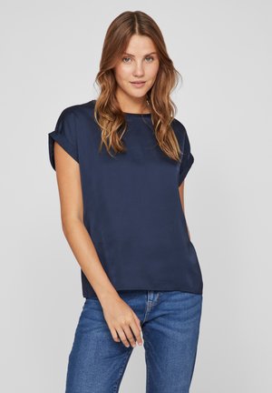 Marineblauwe blouse met korte mouwen en een ronde halslijn. Glad stof, ontspannen pasvorm, opgerolde mouwen. Gecombineerd met blauwe spijkerbroeken, staand op een effen achtergrond.