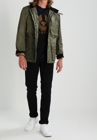 Olivově zelená mikina na zip s několika předními kapsami, černým límcem a kostkovanou podšívkou, v kombinaci s černými kalhotami a bílými teniskami.