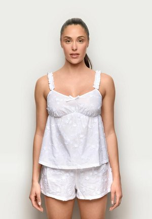 Frau mit zurückgebundenem dunkelbraunem Haar, trägt ein ärmelloses weißes Oberteil mit Blumenstickerei und passende Shorts, steht vor einem schlichten hellen Hintergrund.