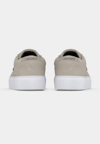 Ζευγάρι μπεζ αθλητικών παπουτσιών Lacoste, εμφανιζόμενα από πίσω με λευκές σόλες και το όνομα της μάρκας στην πίσω ετικέτα, σε λευκό φόντο.