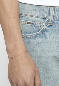 Pulsera de cadena de plata con un acento en forma de estrella. Lleva en una muñeca, combinada con unos jeans de denim azul claro con un acabado descolorido.