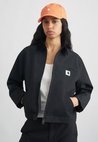 Νεαρή γυναίκα με μακριά σκούρα μαλλιά, φόρεμα πορτοκαλί καπέλο, μαύρο ζακέτα με φερμουάρ πάνω από λευκό tank top, στέκεται με τα χέρια της στις τσέπες της ζακέτας.