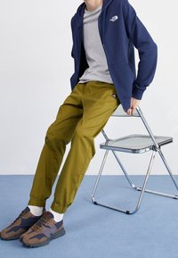 Marinblå zip-up hoodie, grå t-shirt, olivgröna tapered byxor och grå-bruna sneakers med svarta accenter. Sitter på en kromstol.