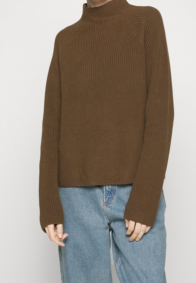 Personne portant un pull en maille côtelée marron à manches longues et un jean bleu denim sur un fond clair uni.