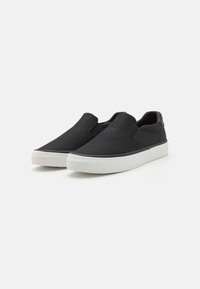 Sorte slip-on sko med et teksturert stoffoverdel, flat hvit gummisåle og elastiske sidepaneler for enkel bruk.