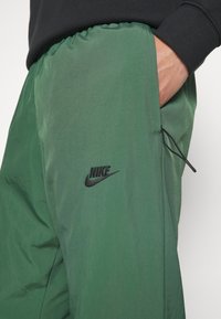 Zelené sportovní kalhoty s tkaničkou v pase, kapsou a černým logem Nike na levém stehně. Hladká, lehká tkanina s jemnou texturou.