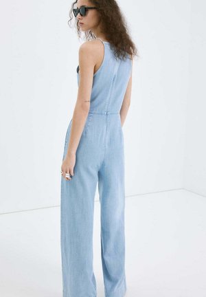 Kobieta o kręconych włosach, nosząca czarne okulary przeciwsłoneczne i jasnoniebieski jeansowy kombinezon bez rękawów, stoi na białym tle.