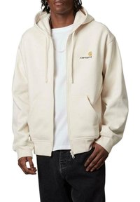 Sudadera cremosa con cremallera y cordones, con un pequeño logotipo amarillo en el lado izquierdo del pecho, combinada con una camisa blanca y jeans oscuros.