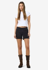 Mladá žena s dlhými rovnými vlasmi má na sebe biele skrátené tričko, čierne riflové šortky a hnedé čižmy po kolená, stojí na bielom pozadí.