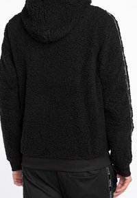 Schwarzer, strukturierter Fleece-Hoodie mit Kapuze, getragen von einer Person, die von hinten zu sehen ist, kombiniert mit schwarzen Hosen mit seitlichen Streifen.