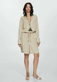 Beige, lightweight, long-sleeve blouse with a button front, paired with matching elastic-waist shorts. Accessories include a triangular necklace.