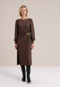 Długa, brązowa sukienka prążkowana z bufkami na rękawach, taliowana, z złotą okrągłą klamrą paska, zestawiona z czarnymi kozakami za kolano.