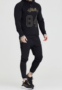 Felpa sportiva nera con accenti dorati, che presenta il numero "84" e il logo. Abbinata a pantaloni sportivi coordinati, anch'essi con dettagli dorati. Sneakers nere.
