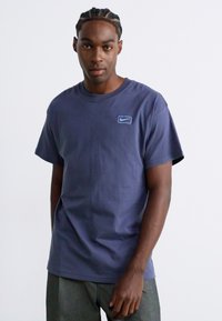 Ung mand med fletninger iført en marinblå Nike t-shirt og grå bukser, stående mod en almindelig hvid baggrund.
