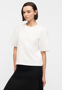 Γυναίκα με κοντά σκούρα μαλλιά, φορώντας λευκή μπλούζα με κοντά μανίκια και ραβδώσεις στην άκρη, καθώς και μαύρη φούστα με ρυτίδες, στέκεται μπροστά σε λευκό απλό φόντο.