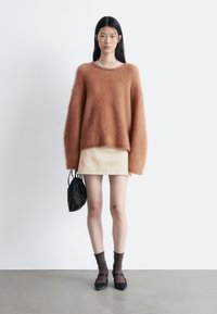 Übergroßer, flauschiger oranger Strickpullover mit langen Ärmeln; kombiniert mit einem beigen Leder-Minirock, einer schwarzen Kordelzugtasche, braunen Socken und schwarzen Ballerinas.