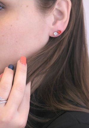 Orecchini a perno con gemme rosse e trasparenti indossati all'orecchio di un modello, che mostrano una finitura lucida. I capelli coprono parzialmente l'orecchio, mentre una mano accarezza delicatamente i capelli.