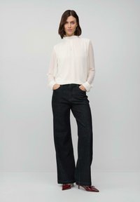 Woman standing with hands in pockets wearing a white high-neck blouse, wide-leg black jeans, and burgundy pointed-toe heels.