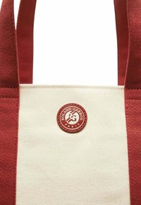 Sac en toile avec des poignées et des accents rouges, corps crème, présentant un logo rouge circulaire avec le texte "Roland Garros" et "Paris".