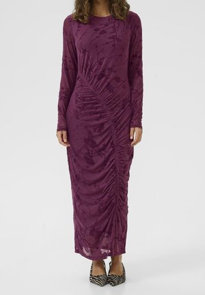 Femme portant une robe longue à manches longues froncées, de couleur violet foncé avec des motifs floraux et des talons pointus à imprimé zèbre.