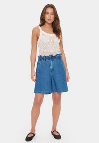 Femeie îmbrăcată cu un top alb din croșetat fără mâneci, pantaloni scurți din denim albastru cu talie înaltă, largi și pantofi Mary Jane negri, stând pe un fundal alb.