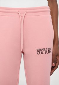 Roosa dressipükste komplekt, mis on valmistatud pehmest kangast, elastse vöökoha ja nööpidega, millel on must tikand "VERSACE JEANS COUTURE" logo detail.