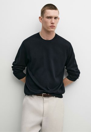 Junger Mann mit kurzen Haaren, der ein schwarzes Langarmshirt und beigefarbene Hose mit einem braunen geflochtenen Gürtel trägt, steht vor einem schlichten weißen Hintergrund.