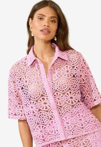 Rozā mežģīņu krekls ar ziedu ģeometrisko rakstu, īsām piedurknēm un apkakles dekoltē, ar pogu detaļām un puscaurspīdīgu faktūru.