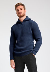 Maglione lavorato a maglia blu navy con colletto alto e zip a metà, con texture a coste e maniche aderenti. Indossato con pantaloni scuri e strutturati.
