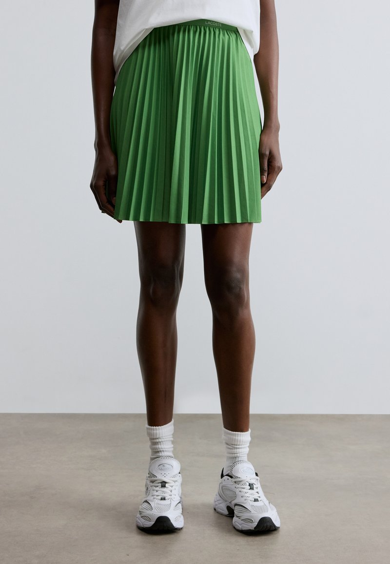 Πράσινη φούστα με πιέτες και ελαστική μέση, σε συνδυασμό με λευκά αθλητικά παπούτσια και λευκές κάλτσες αστραγάλου. Απλός σχεδιασμός, χωρίς εμφανή σχέδια ή διακοσμήσεις.