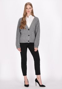 Cardigan gri tricotat cu decolteu în V, șase nasturi negri și manșete cu nervuri, asortat cu pantaloni negri ajustați și pantofi cu tocuri înalte negri.