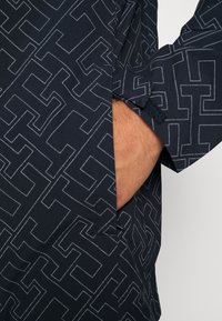 Námořnická košile s geometrickým vzorem v kontrastní světle šedé barvě. Má boční kapsu a zapínání manžety na suchý zip. Hladká textura.