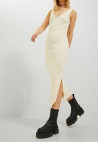 Frau in einem ärmellosen, cremefarbenen Strick-Midikleid mit Seitenschlitz, kombiniert mit schwarzen klobigen Plateaustiefeln und einer dünnen schwarzen Choker-Halskette.