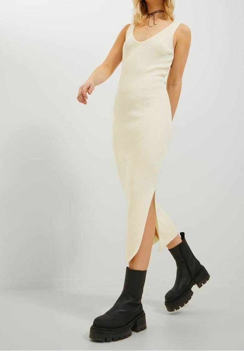 Vrouw in een mouwloze crèmekleurige gebreide midi-jurk met zijsplit, gecombineerd met zwarte chunky platformboots en een dunne zwarte choker.