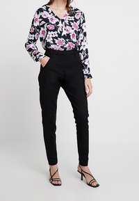 Kvinna iklädd svarta åtsittande byxor, blommig svartvit blus med rosa blommor, och svarta sandaler med klackar och remmar, stående med en hand i fickan.